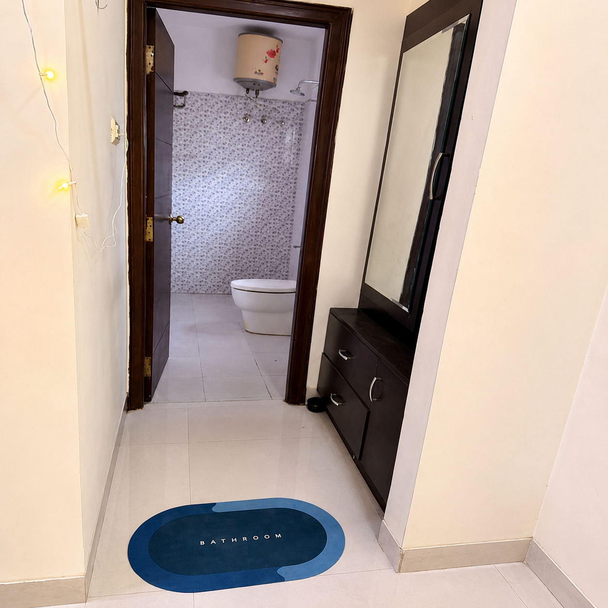 Bathroom, bathroom interior, modern bathroom, compact bathroom, bathroom entrance, bathroom hallway, bathroom design, contemporary bathroom, clean bathroom, tiled bathroom, patterned wall tiles, white bathroom tiles, glossy floor tiles, bathroom toilet, modern toilet, bathroom water heater, wall mounted water heater, bathroom mirror, dressing mirror, vanity cabinet, dark wood vanity, bathroom storage cabinet, bathroom doorway, wooden door, open bathroom door, narrow hallway, apartment bathroom, residential bathroom, well maintained bathroom, bright bathroom, indoor lighting, warm string lights, bathroom floor mat, blue bathroom mat, clean floor, practical bathroom, functional bathroom, compact home bathroom, student-friendly bathroom, student accommodation bathroom, shared accommodation bathroom, student living space, business professional bathroom, professional accommodation, convenient bathroom, urban apartment bathroom, rental property bathroom, modern home interior, real estate bathroom, comfortable living space, organized bathroom.
